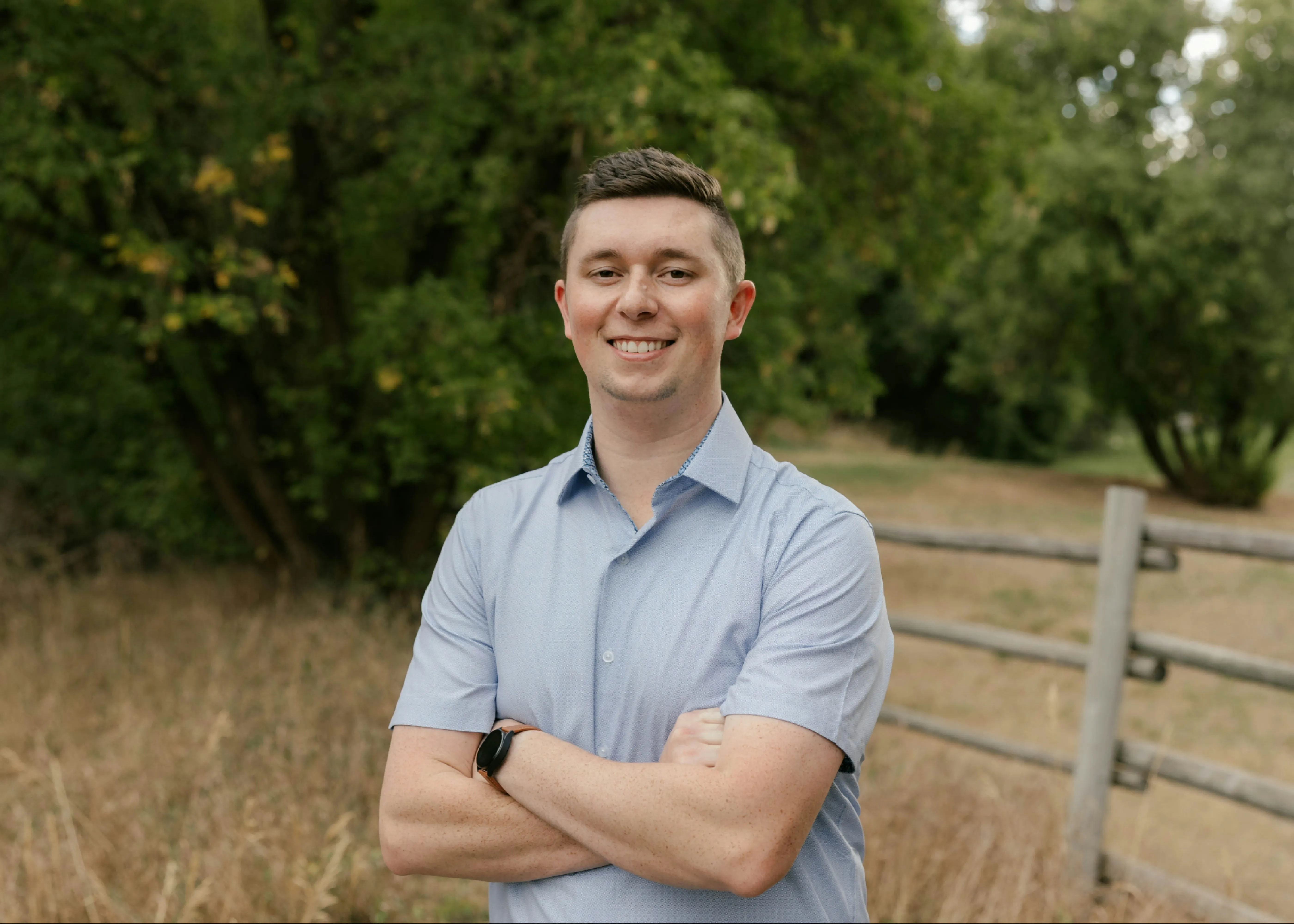 Jace Cox standing outdoors beside a split-rail fence in late summer, arms folded.