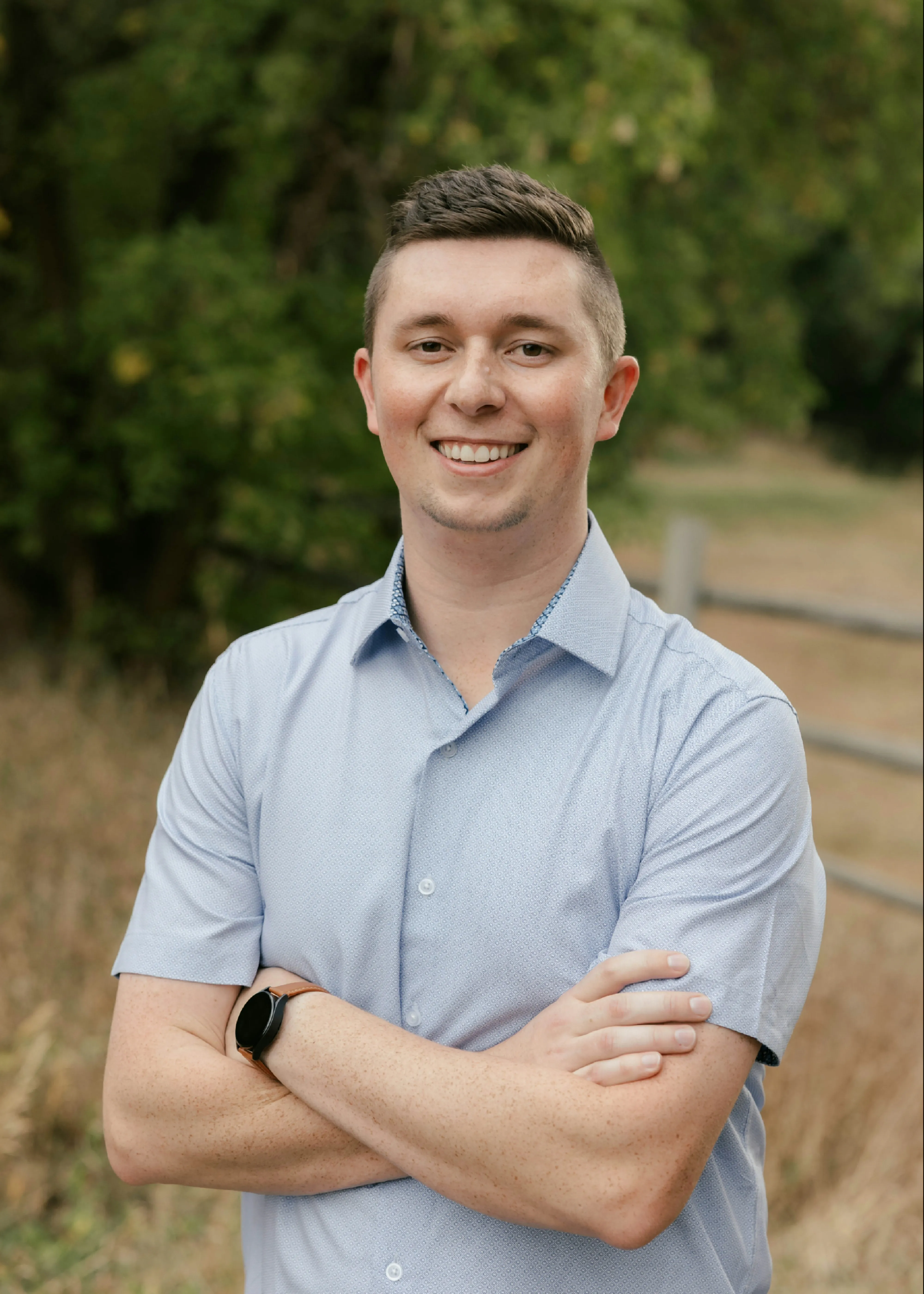 Jace Cox outdoors in a light blue shirt, arms folded, smiling.